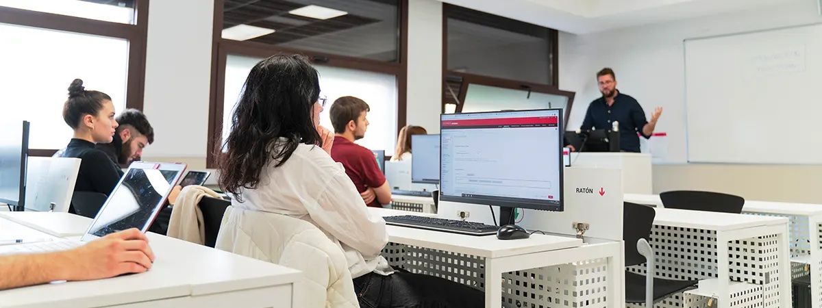 Un aula moderna con estudiantes atentos mientras un profesor explica delante de una pizarra.