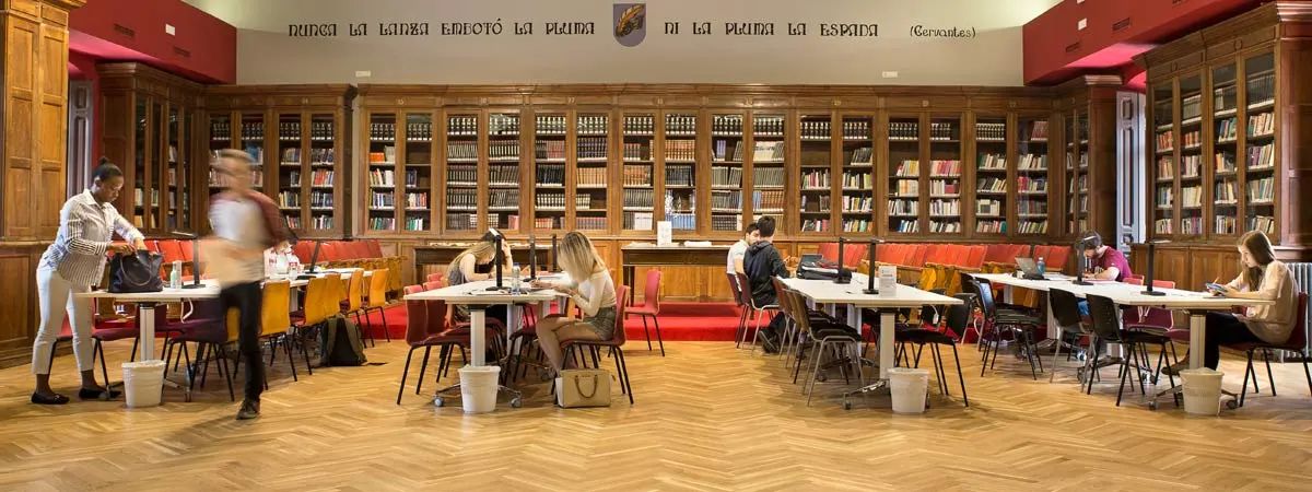 Una vista interior de una biblioteca con mesas de estudio y estanterías llenas de libros.