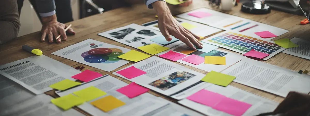 Sobre una mesa se encuentran documentos, gráficos y notas adhesivas de colores, mientras una mano señala un papel.
