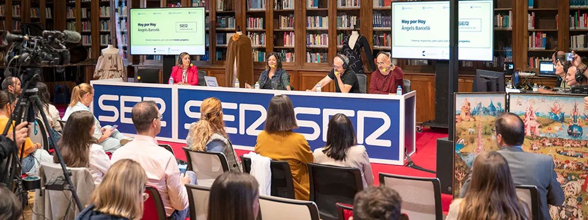 Una mesa redonda con presentadores y una audiencia atenta en un entorno de biblioteca.