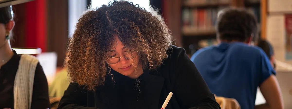 Una persona con cabello rizado y gafas escribe en una mesa en un entorno de biblioteca.