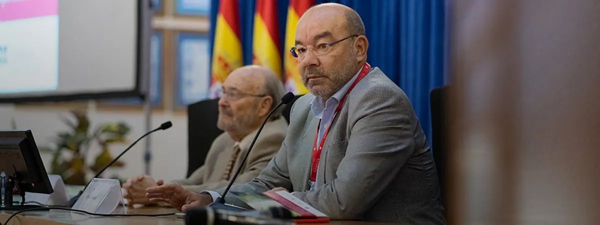 Dos hombres están sentados en una mesa durante una conferencia, con banderas de España al fondo.