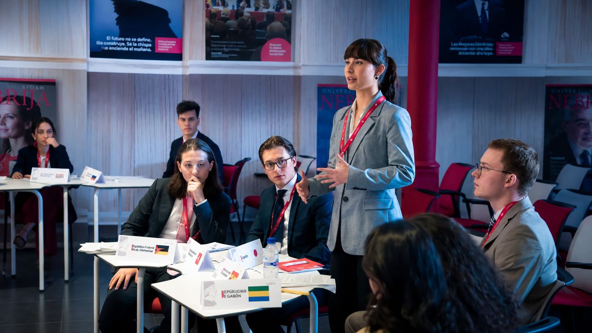 Un grupo de jóvenes participan en una discusión, donde una mujer está de pie presentando su punto de vista.