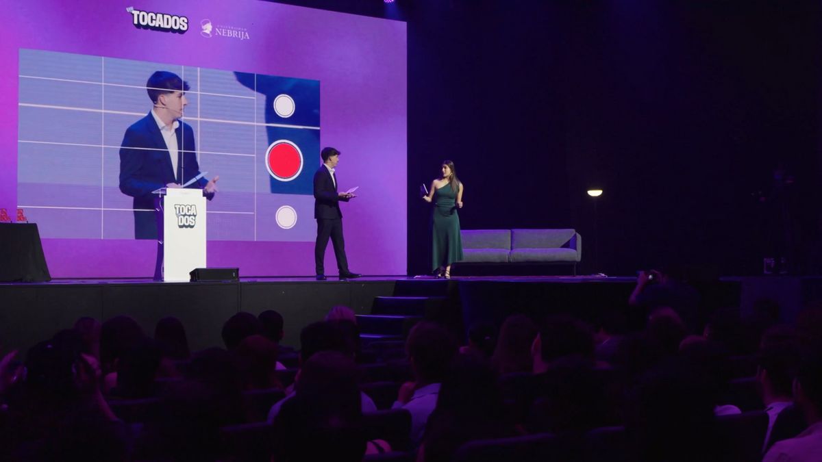 Un hombre y una mujer están en el escenario de un evento, interactuando frente a una presentación visual con elementos gráficos.