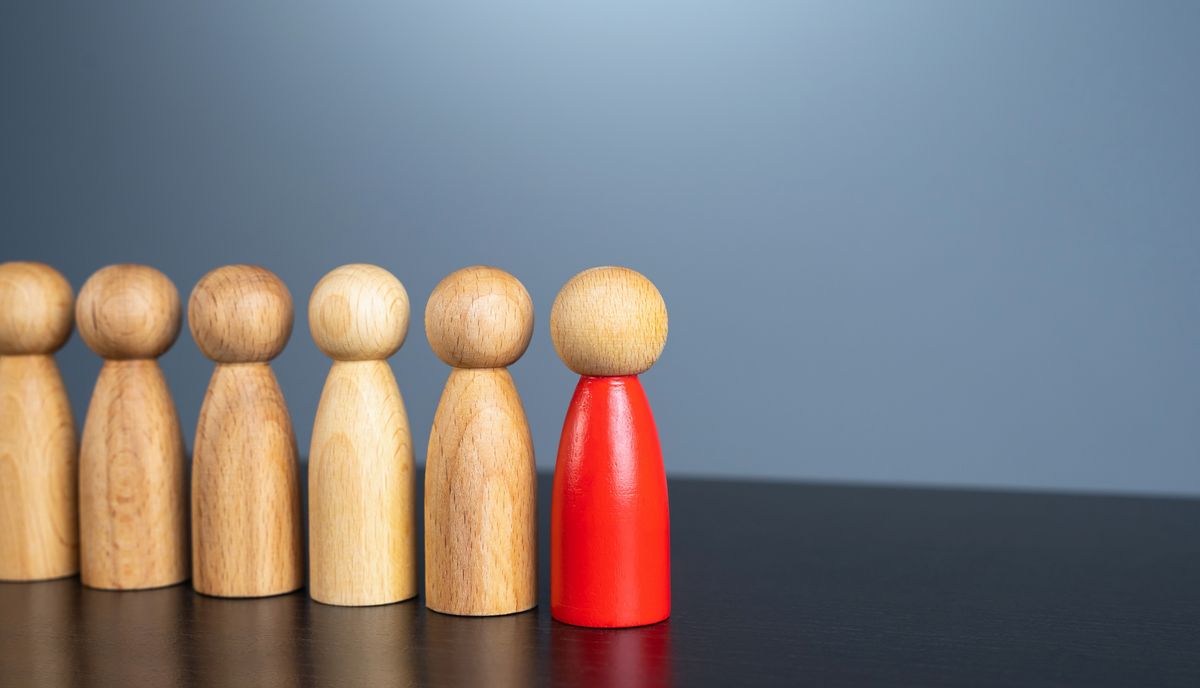 La imagen muestra una fila de figuras de madera, con una figura roja destacando entre las demás de color natural.