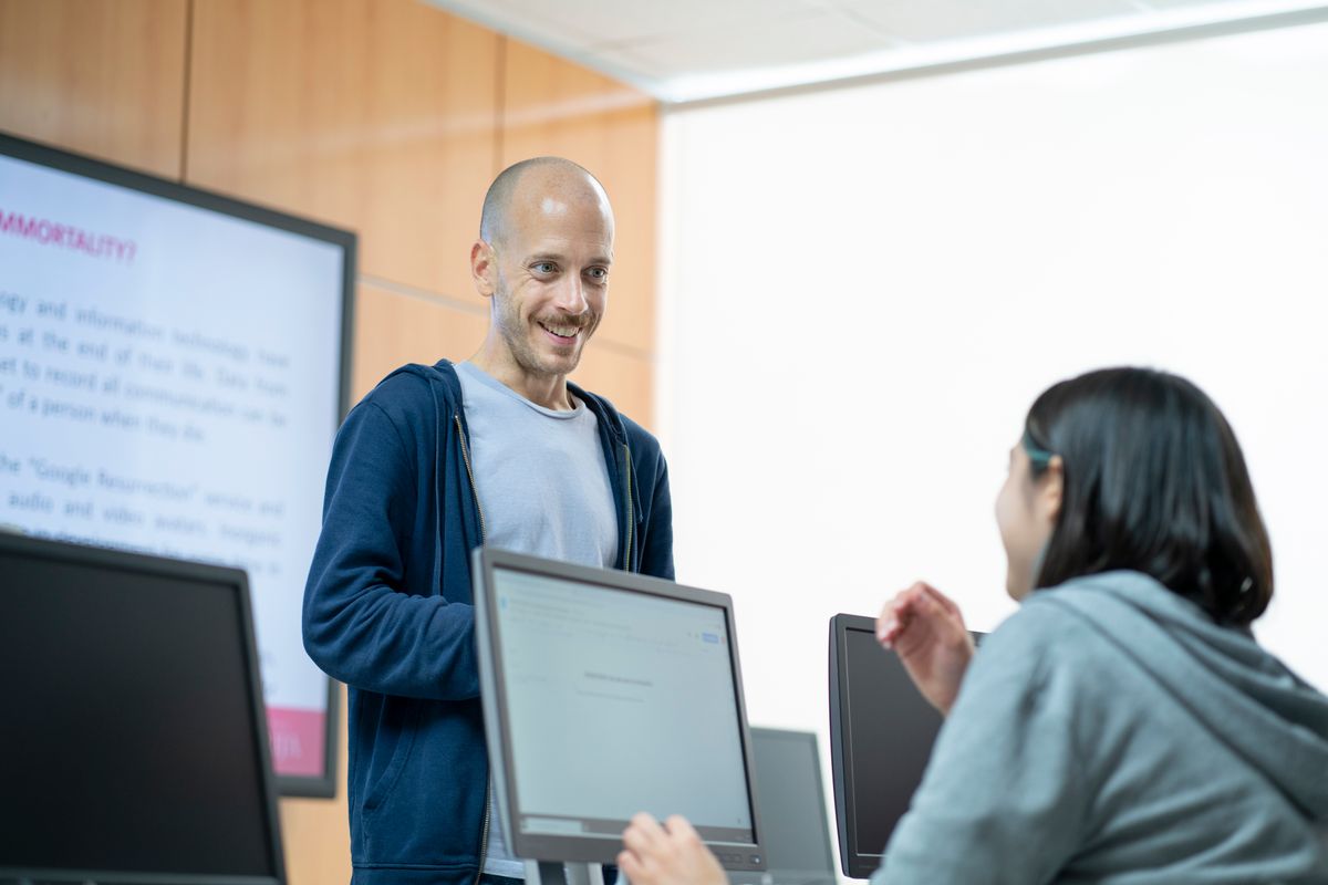 Un hombre sonriente está conversando con una mujer en un aula frente a computadoras.