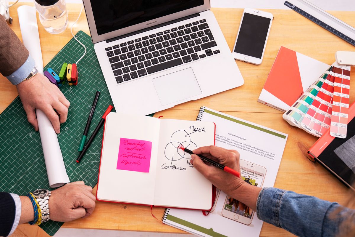En la mesa se observa un ambiente de trabajo colaborativo con personas tomando notas y usando tecnología.