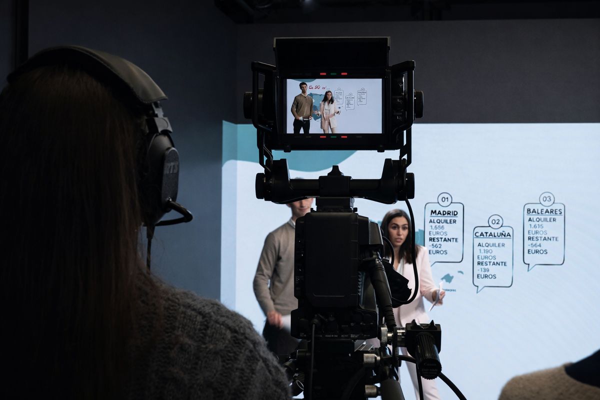 La imagen muestra a dos presentadores de pie frente a una pantalla con información sobre precios en diferentes regiones de España, mientras una cámara graba la presentación.