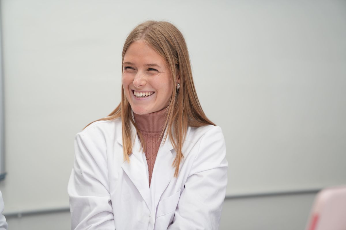 Una joven sonriente vestida con una bata de laboratorio blanca en un entorno académico.