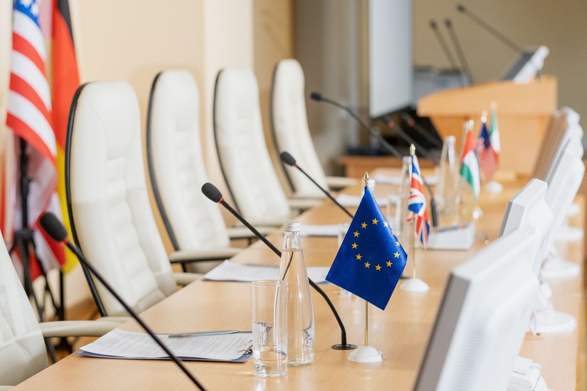 La imagen muestra una sala de reuniones con varias sillas y banderas de diferentes países, destacando la bandera de la Unión Europea.