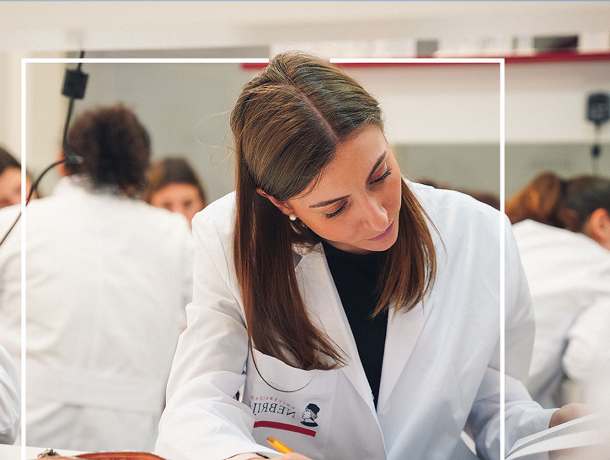 Una mujer joven en un laboratorio, vestida con bata blanca, escribiendo concentradamente en una hoja de papel.