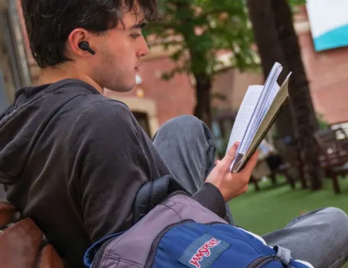 Un joven sentado en un parque, leyendo un cuaderno mientras escucha música con auriculares.