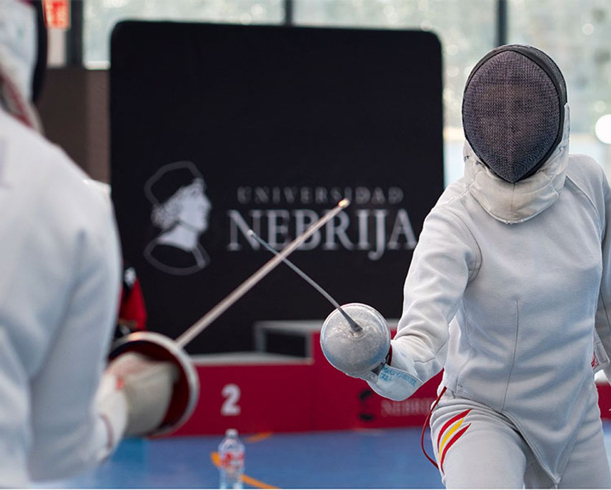 Dos esgrimistas compiten en un torneo con la bandera de la universidad Nebrija de fondo.