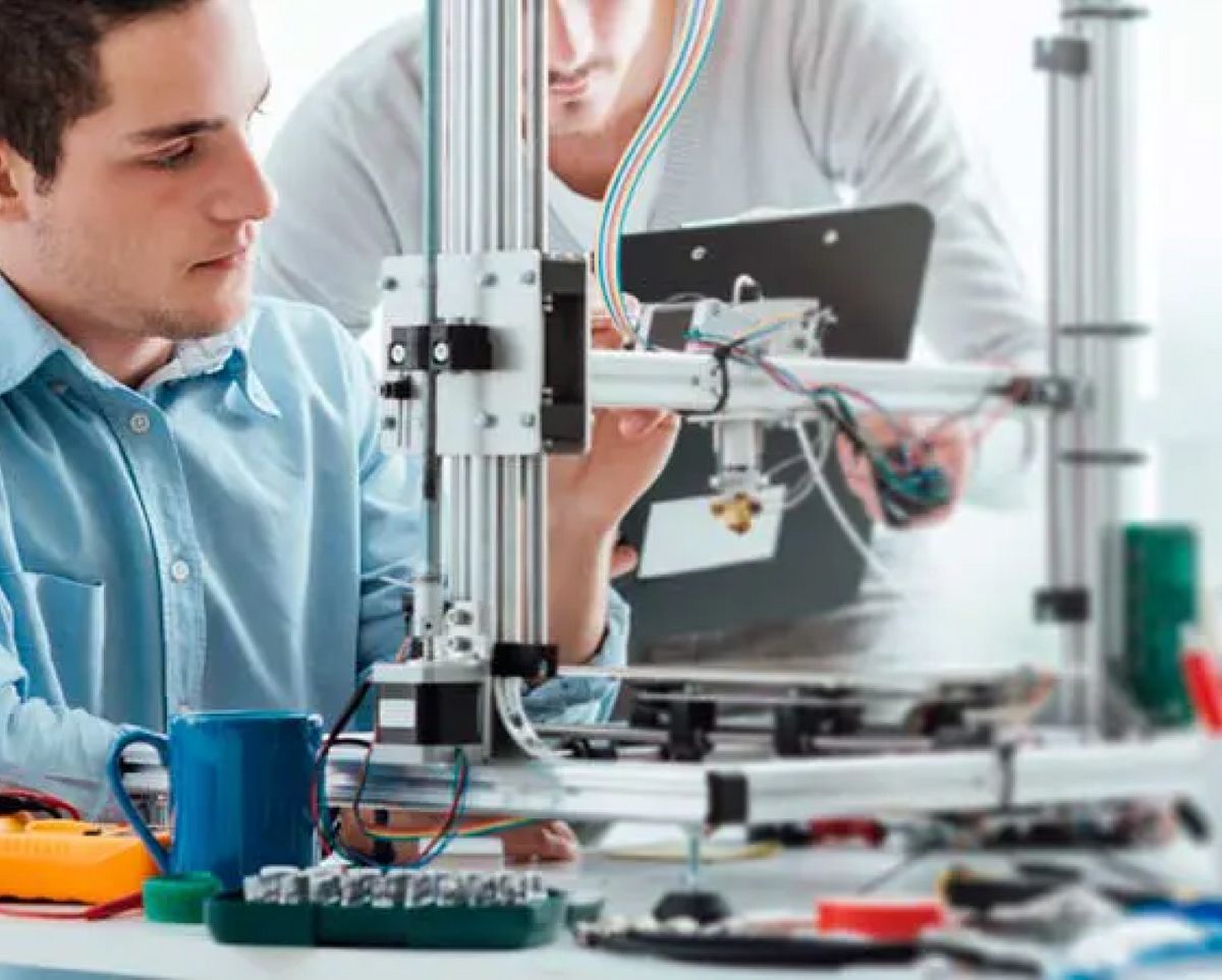 Un hombre trabaja en una impresora 3D mientras otro observa y sostiene una tablet.