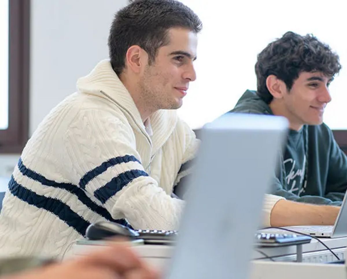 Dos jóvenes estudiantes están sentados en una clase, uno de ellos sonríe mientras trabaja en su computadora portátil.