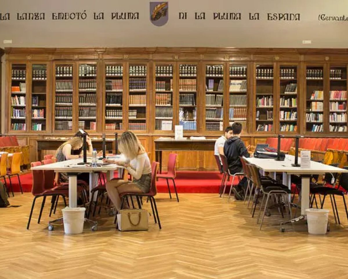 Una biblioteca con varios estudiantes trabajando en mesas rodeados de estanterías llenas de libros.