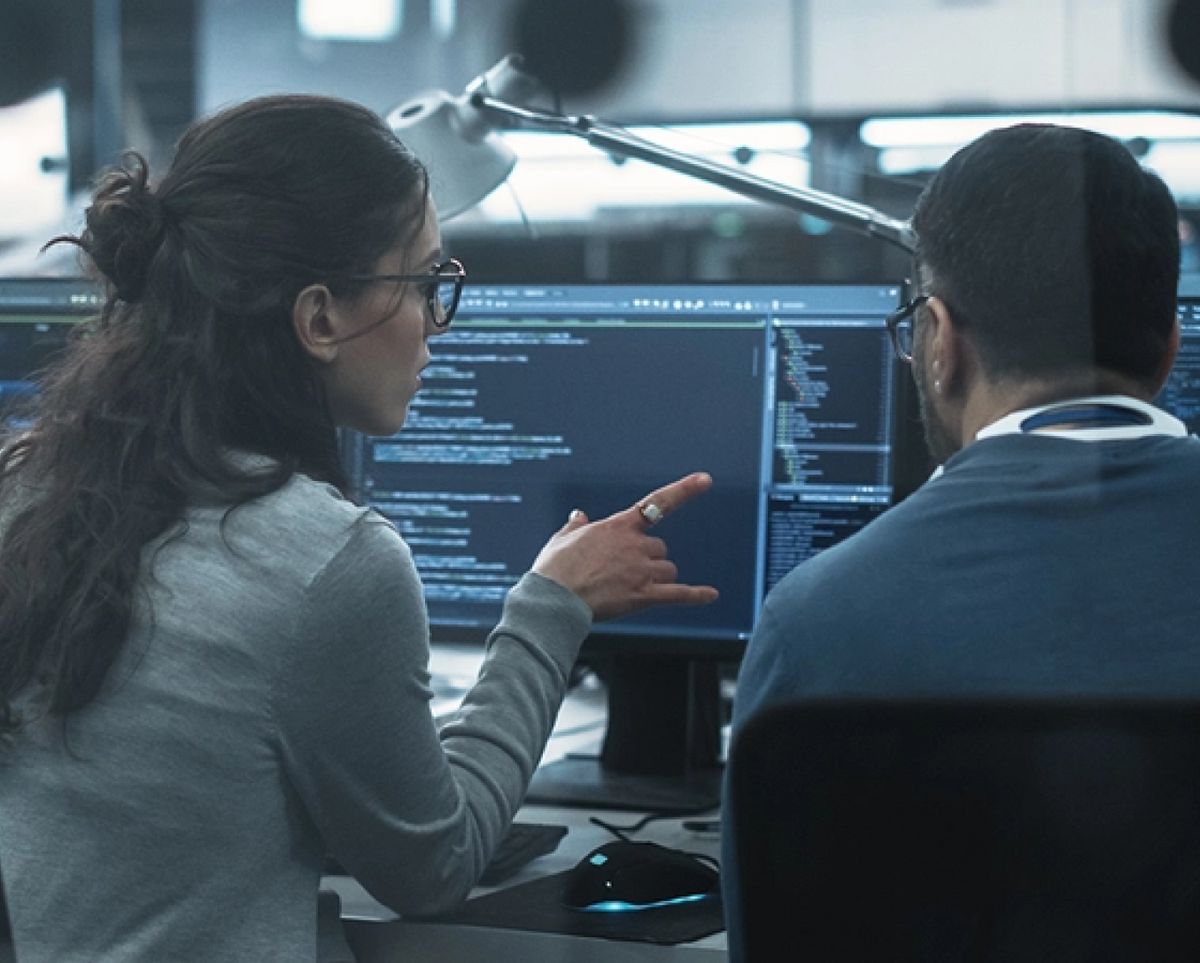 Dos personas conversan y trabajan en programación frente a una computadora con código en la pantalla.