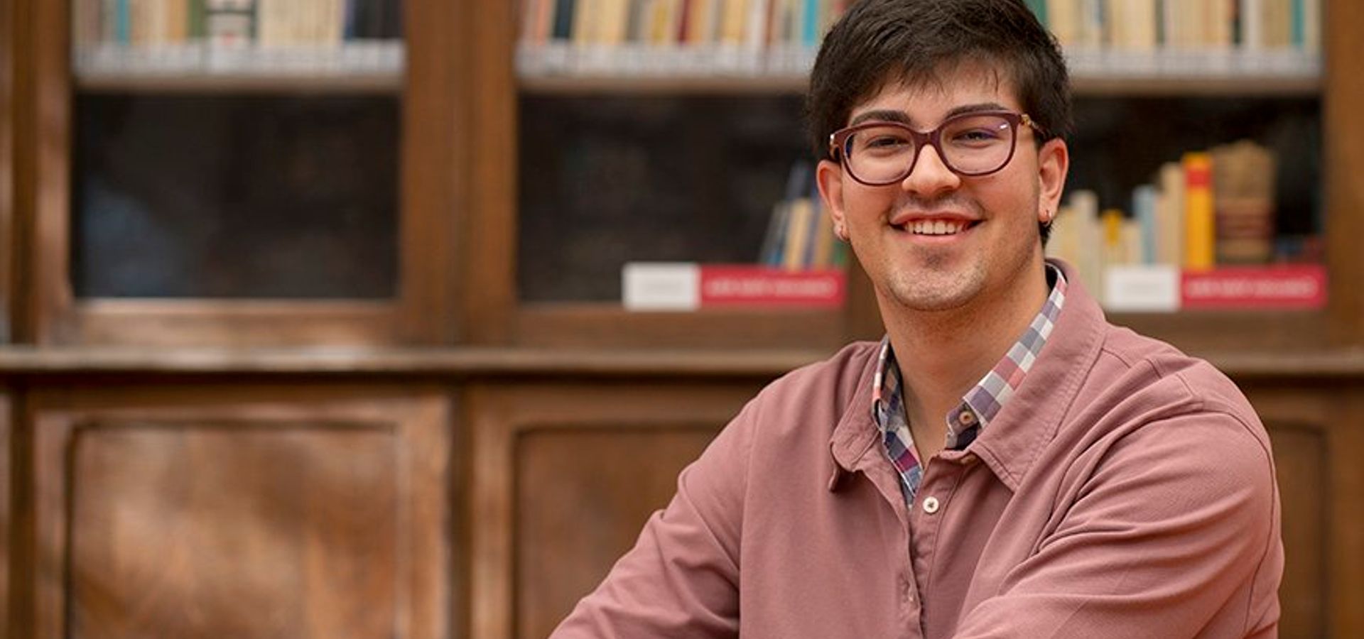 Un joven sonriente sentado en una mesa de una biblioteca con estanterías llenas de libros al fondo.