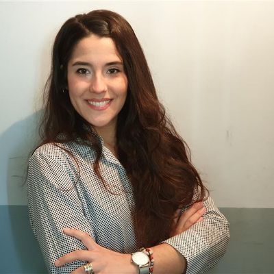 Una joven sonriente con cabello largo y oscuro está de pie con los brazos cruzados frente a una pared clara.