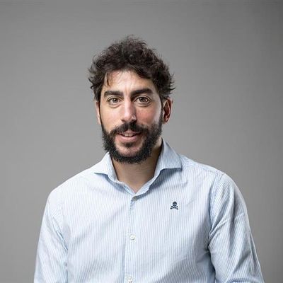 Un hombre sonriente con barba y cabello rizado, vestido con una camisa de rayas lisas en un fondo gris.