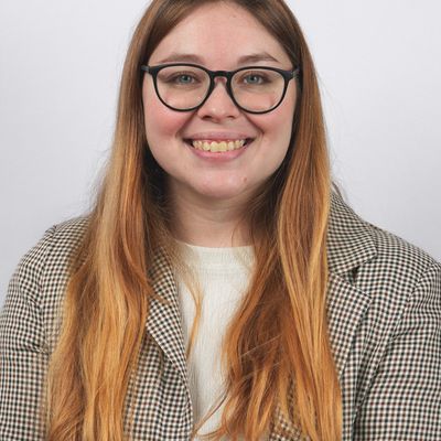 Una mujer sonriente con cabello largo y lacio, vestida con una chaqueta a cuadros y gafas.