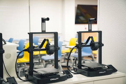 La imagen muestra dos impresoras 3D en un entorno de aula con sillas de colores.