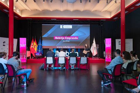 La imagen muestra un seminario titulado 'Nebrija Emprende' con una presentadora frente a un panel y una pantalla al fondo.