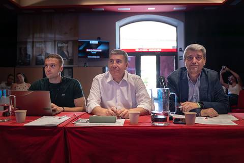 Tres hombres sentados detrás de una mesa en un ambiente formal, observando una presentación.