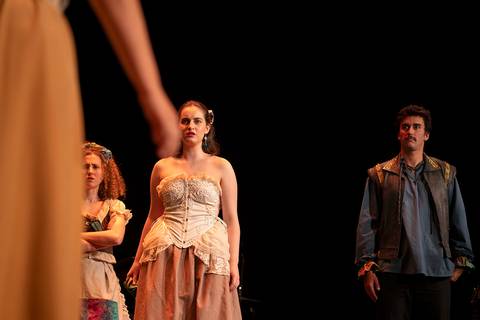 Un grupo de actores en una escena teatral, donde una mujer vestida de manera elegante es el centro de atención.