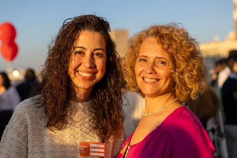 Dos mujeres sonrientes posan juntas al aire libre durante un evento social al atardecer.