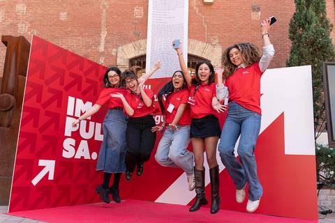 Un grupo de cinco mujeres sonrientes y entusiastas saltando frente a un fondo rojo con un mensaje inspirador.