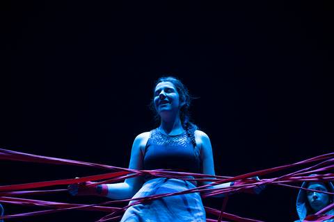 Una joven rodeada de cintas rojas, con una expresión de alegría en un escenario oscuro.