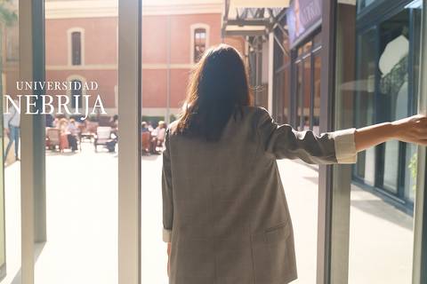 Una persona entra en un edificio universitario acompañado de un ambiente soleado y un grupo de gente en el exterior.