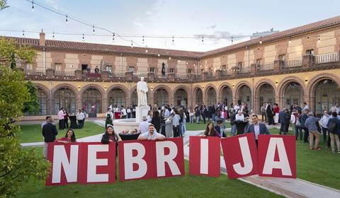La imagen muestra un evento al aire libre en un patio con personas reunidas y un grupo sosteniendo un letrero grande que dice 'NEBRIJA'.
