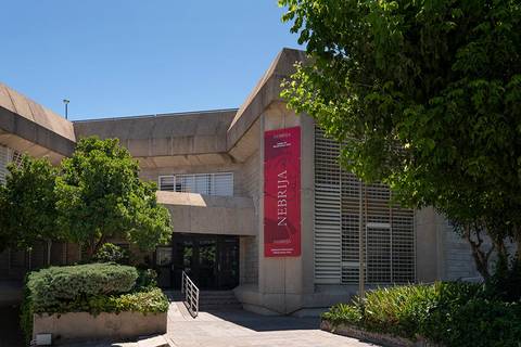La imagen muestra un edificio moderno con un banner que dice 'NEBRIJA' y una vegetación exuberante alrededor.