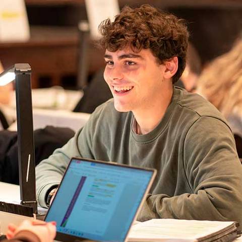 Un joven sonriente trabaja en su computadora mientras está rodeado de otras personas.