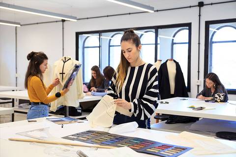 Un grupo de estudiantes trabaja en un estudio de diseño de moda, con patrones y telas en diversas mesas.