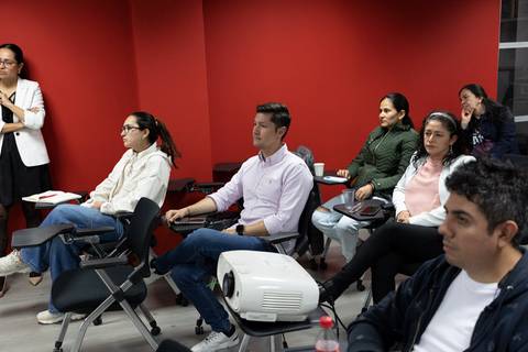 Un grupo de personas sentadas en un aula con una pared roja de fondo.