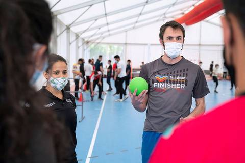 Un joven con una pelota verde sostiene una charla en un evento de triatlón mientras lleva una camiseta que dice 'FINISHER'.