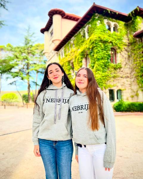 Dos jóvenes vistiendo sudaderas con el logo de Nebrija posan frente a un edificio rodeado de vegetación.