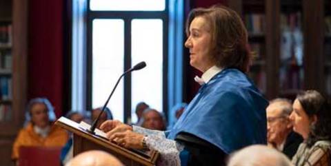 Una mujer con toga se encuentra en un atril hablando frente a un público en un salón.