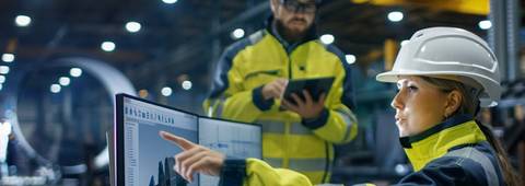 Una mujer con casco y chaqueta de seguridad señala la pantalla de una computadora mientras un hombre con bagaje similar observa.