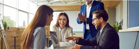 Un grupo de empleados sonríe mientras interactúan alrededor de una computadora portátil en una oficina moderna.