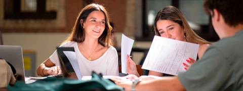 Dos estudiantes revisan documentos mientras estudian juntas en un entorno luminoso.