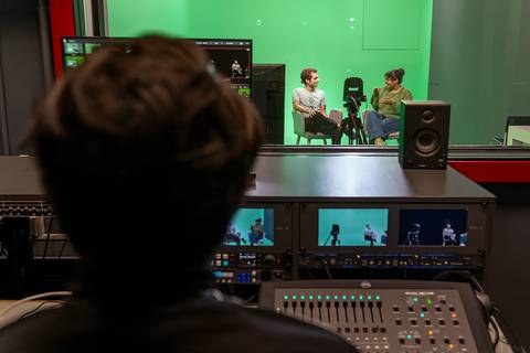 Un grupo de personas está entrevistando en un estudio con fondo verde mientras alguien controla la grabación desde la consola.