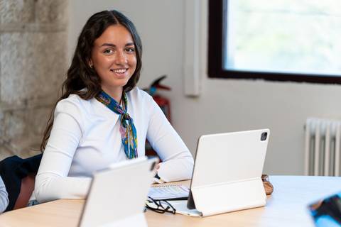 Una joven sonriente se sienta en una mesa de trabajo con una tablet frente a ella.