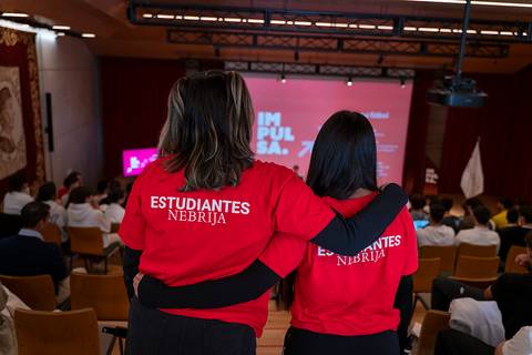 Dos estudiantes con camisetas rojas que dicen 'ESTUDIANTES NEBRIJA' observan un evento desde el público.