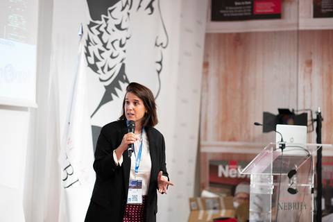 Una mujer está dando una presentación en un evento, con un micrófono en la mano y una pantalla detrás.