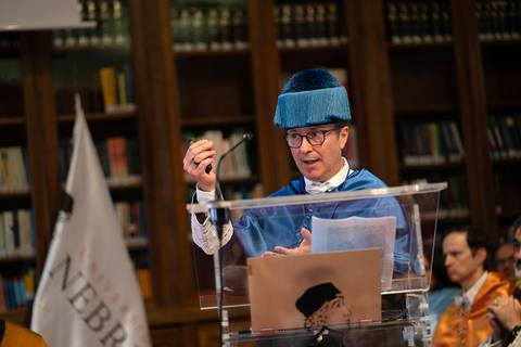 Una persona vestida de toga y birrete está hablando en un podio frente a un público en una biblioteca.