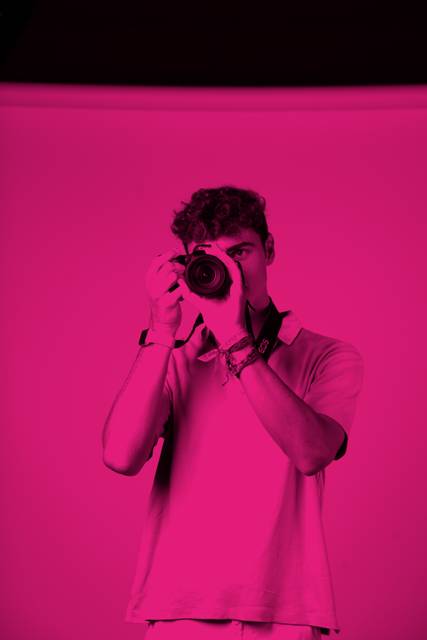 Un joven posando con una cámara en un fondo rosa.